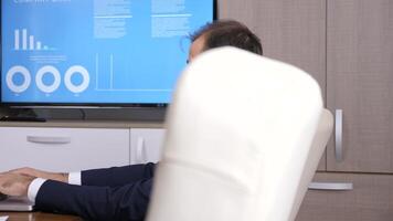 Revealing shot of businessman working on the computer in conference room. There is a TV with animated data and graphs in the background. Dolly slider 4K footage video