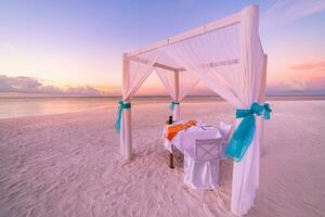 Romantic dinner setting on the beach at sunset sky. Honeymoon or anniversary couple destination dining. Romance over white sand and endless sea view. Colorful love sunset skyscape photo