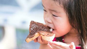 Slice of bread covered with chocolate cream is being eaten by kid girl with gusto. Child are hungry. Children eat snacks in afternoon. Empty space to enter text. photo