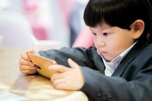 Asian boy watching cartoons with his smartphone. A 3 year old child wears black suit. Concept of baby use of digital media in long time. photo