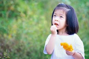 Cute Asian kid girl enjoying battered fried papaya as snack during day. Children try to eat local food from nature. Empty space for entering text. 4 year old child wears white t shirt. photo