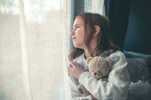 a pensive cute girl sits and looks out the window, hugging a teddy bear. photo