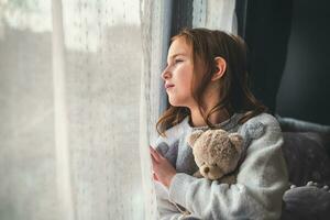 a pensive cute girl sits and looks out the window, hugging a teddy bear. photo