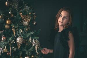 linda pequeño niña siguiente a Navidad árbol. Navidad, nuevo año, oscuro tonos foto