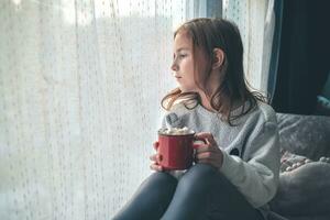 a cute girl on the windowsill holds a red mug of cocoa and marshmallow and looks out the window. photo