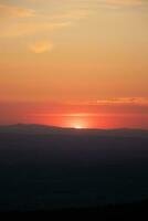 Disc of sunset with reddish hues sets behind the mountains in the distance. Blue, orange and golden sky. Airplane flying over in red sun. Abstract sky photo