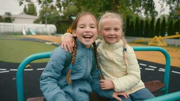 twee weinig meisjes vrienden zussen rijden schommel lachend glimlachen vreugde geluk kinderjaren speelplaats knuffel pret schoolmeisjes wandelen Speel buiten stad kinderen klasgenoten kinderen samen school- onderwijs aan het leren video