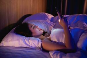 Asian woman using mobile phone smartphone laying on the bed in the bedroom. Sleepy exhausted, can not sleep. Insomnia, addiction concept. Women scrolling social networks on mobile dark bedroom. photo