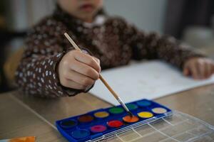 cerca arriba mano de linda pequeño asiático bebé sonriente pintura con vistoso pinturas utilizando acuarela. asiático niña utilizando Cepillo de pintura dibujo color. bebé actividad estilo de vida concepto. foto