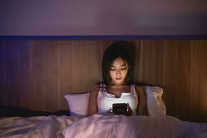 asiático mujer utilizando móvil teléfono teléfono inteligente tendido en el cama en el dormitorio. soñoliento exhausto, lata no dormir. insomnio, adiccion concepto. mujer desplazamiento social redes en móvil oscuro dormitorio. foto