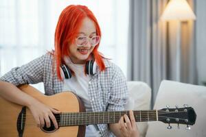 Asian woman wearing glasses and headphone playing guitar while sitting on sofa in the living room at home. Asian women writing song while playing guitar at home. Compose song music concept. photo