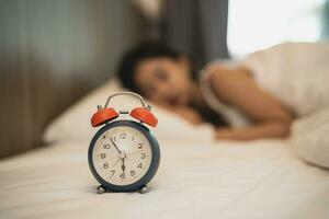 cerca arriba alarma reloj. mujer dormido en cómodo cama con sedoso ropa de cama en el Mañana. mujer acostado en cama y acuerdo ojos cerrado mientras cubierto con frazada. alegre y cómodo. foto