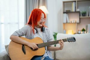 asiático mujer vistiendo lentes y auricular jugando guitarra mientras sentado en sofá en el vivo habitación a hogar. asiático mujer escritura canción mientras jugando guitarra a hogar. componer canción música concepto. foto