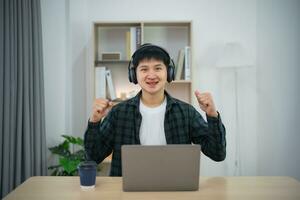 Smart Asian man wearing headphone with braces smiling raise hand cheerful working with computer laptop. Work form home. New normal social distancing lifestyle. Work form anywhere concept. photo