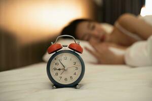 cerca arriba alarma reloj. mujer dormido en cómodo cama con sedoso ropa de cama en el Mañana. mujer acostado en cama y acuerdo ojos cerrado mientras cubierto con frazada. alegre y cómodo. foto