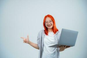 Teen woman wearing glasses cheerful and point finger side and holding laptop isolated on white or grey background, women using laptop to search or working or typing. Technology education concept. photo