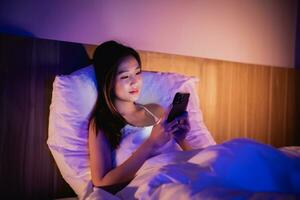 Asian woman using mobile phone smartphone laying on the bed in the bedroom. Women scrolling social networks on mobile. Content female lying in bed under blanket and watching movie on smartphone. photo