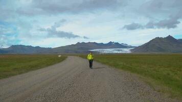 Female tourist with backpack goes by dirt road on background of mountains and glacier in Iceland. Freedom and travel concept. 4K video