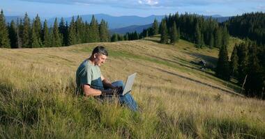 uma homem senta em uma lindo Prado dentro a montanhas, trabalho em uma computador portátil. conceito do freelancer, digital nômade ou controlo remoto escritório. video