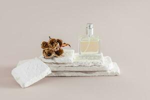 A clear bottle of women's perfume or cosmetic spray stands on a stack of white brick slices with a dried flower. Front view. Product presentation. photo