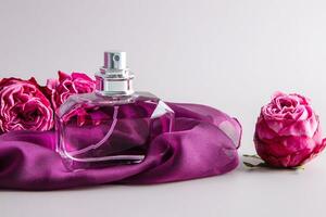 A chic bottle of women's perfume stands on a chiffon purple scarf among fresh roses. Expensive fragrance. Front view. Pastel background. photo