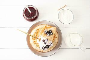 A large plate of freshly made crepes, sour cream, a jar of jam, milk in a jug on a white wooden table. Top view. Delicious breakfast. photo