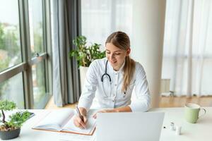 Young professional female doctor wearing uniform taking notes in medical journal, filling documents, patient illness history, looking at laptop screen, student watching webinar photo