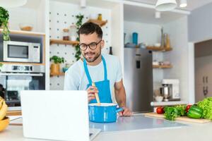 un joven hombre gasta un día a hogar, prepara comida de vegetales en el cocina, un hombre en hogar ropa y con un barba, usos un ordenador portátil a aprender en línea Cocinando foto