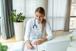 Doctor professional female doctor wearing uniform taking notes in medical journal, filling documents, patient illness history, looking at laptop screen, student watching webinar photo