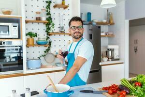 hogar cocinar. soltero hombre en delantal mirando a el ventana, Bebiendo vino desde un vaso mientras Cocinando en el cocina, Cocinando a hogar foto