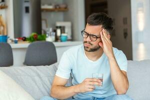 Young man suffering from strong headache or migraine sitting with glass of water on the sofa, millennial guy feeling intoxication and pain touching aching head, morning after hangover concept photo