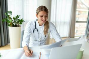 Smiling professional female doctor wearing uniform taking notes in medical journal, filling documents, patient illness history, looking at laptop screen, student watching webinar photo