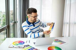 web designer working from home, sitting at table with modern laptop, looking at color samples, happy man freelancer enjoying his remote job, copy space photo