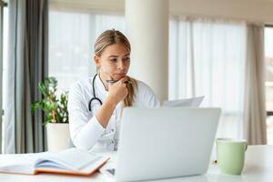 Doctor professional female doctor wearing uniform taking notes in medical journal, filling documents, patient illness history, looking at laptop screen, student watching webinar photo