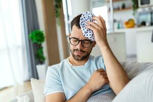 When you hit shelf with your head. Frustrated young man holding ice bag on his head while lying on the couch at home photo