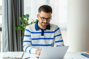 sonriente hombre en lentes sentar a escritorio en oficina hojeada inalámbrico Internet en ordenador portátil dispositivo, contento masculino trabajador relajarse a trabajo descanso foto