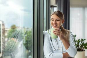Shot of female doctor holding cup of coffee while looking forward window standing in the consultation. Smiling young woman doctor in white medical uniform and stethoscope look in distance planning. photo