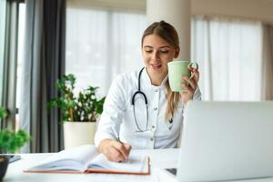 Young professional female doctor wearing uniform taking notes in medical journal, filling documents, patient illness history, looking at laptop screen, student watching webinar photo