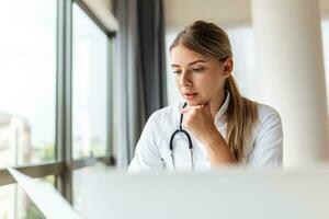 Smiling professional female doctor wearing uniform taking notes in medical journal, filling documents, patient illness history, looking at laptop screen, student watching webinar photo