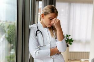 Young doctor looking distressed. Tired exhausted female doctor in uniform at hospital holding her head. Depressed sad doctor feels burnout stress photo