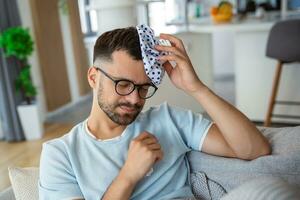 Man feeling bad, sitting on the sofa with headache and applying ice bag photo