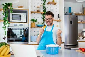 Funny young man singing song into whisk, cooking in modern kitchen, happy male holding kitchenware as microphone, listening to music, dancing, doing housework at home, preparing breakfast photo