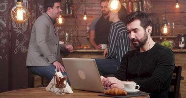 lento diapositiva tiro di un' giovane uomo rallegrare al di sopra di un realizzazione mentre freelance nel un' caffè negozio. Lavorando su il suo il computer portatile con il cuffia Su. video