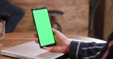 A young womans hand holding a smartphone with green screen. Bar counter in the background. Watching a video on the small screen.