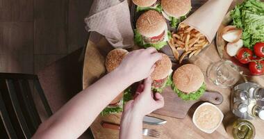 oben Aussicht Komposition mit Burger und ein Köche Hände setzt Sie auf Tisch. viele Burger, Cheeseburger auf ein hölzern Tisch. Französisch Fritten, Gurken, Tomaten und Zwiebeln. video