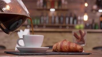 Close up shot of a coffee pot filling a cup to serve with croissant. Blured background of a cozy, warm bar. Vintage wooden decor. video