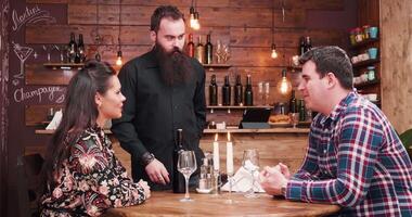 Bearded hipster waiter opening a bottle of wine at the client desk. They are in a vintage rustic pub or restaurant video