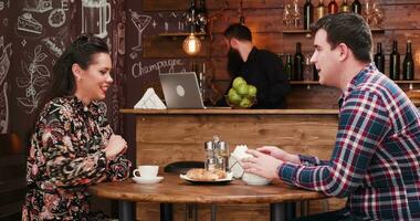 hermosa Pareja comiendo croissants y Bebiendo café en elegante café tienda restaurante pub. un barbado camarero es en el fondo a el mostrador trabajando video