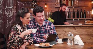 Couple using a digital tablet PC in vintage coffee shop with wooden walls and rustic interior. There is a bearded bartender working in the background at the counter video