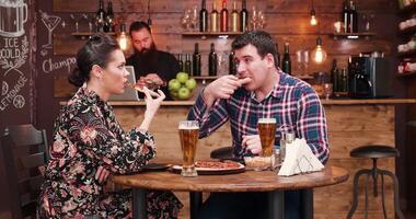 Caucasian couple eating pizza and drinking beer in very cozy and stylish pub or restaurant. A bearded bartender is sitting in the background and works at the counter video